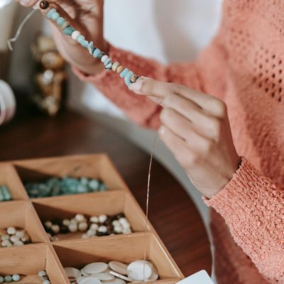 Crop unrecognizable craftswoman in casual clothes sitting at table with box for craftwork and demonstrating partly finished colorful decorative handmade accessory